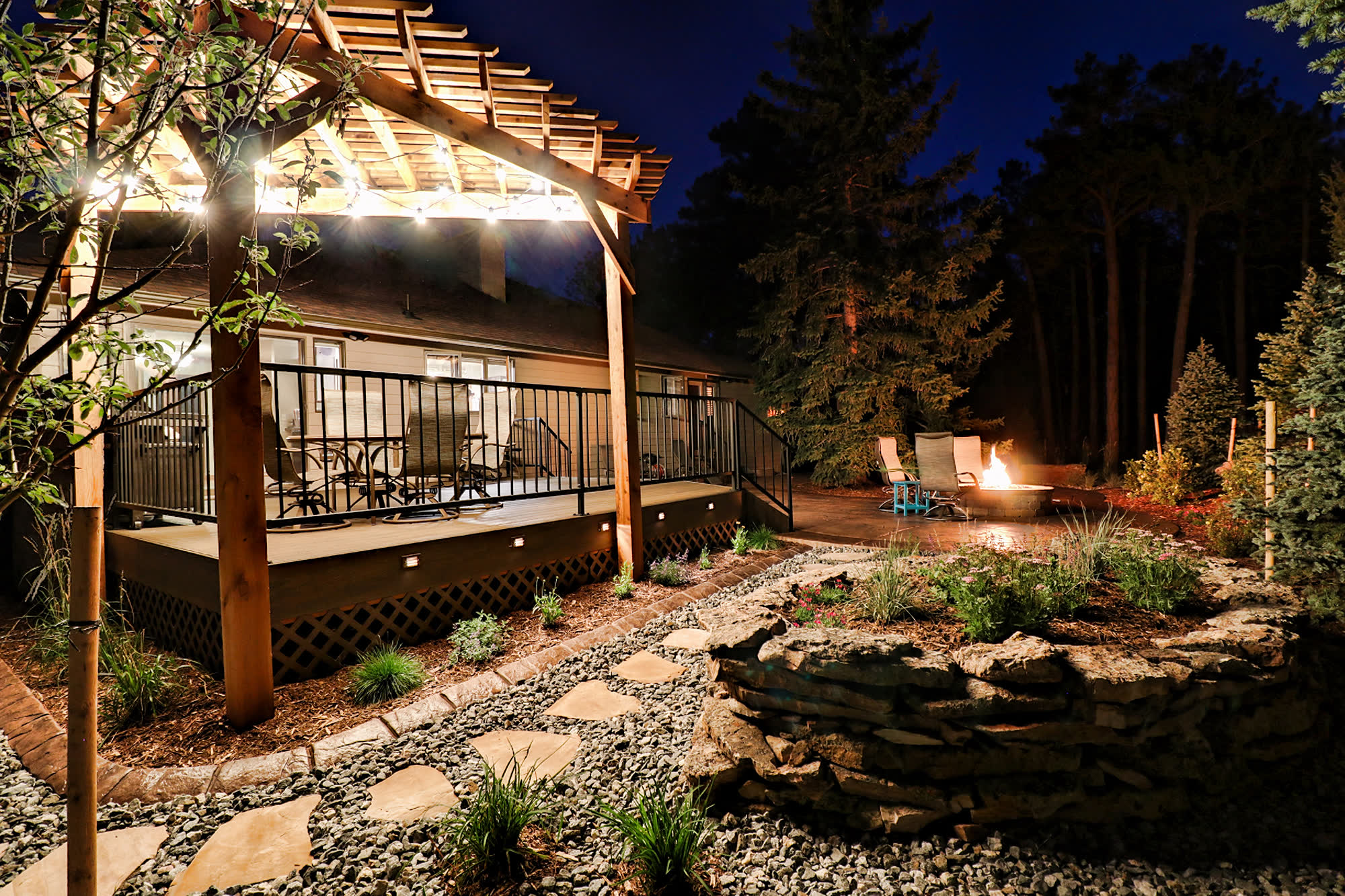 A string-lit cedar pergola over a deck at dusk, with a lit fire pit beyond, at the Amdahl residence